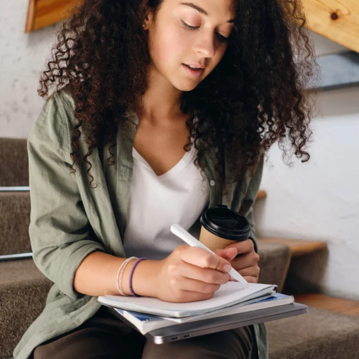 03-NewPatientForms_young-girl-with-dark-curly-hair-sitting-stairs-university-with-cup-coffee-go-hand-thoughtfully-writing-notebook